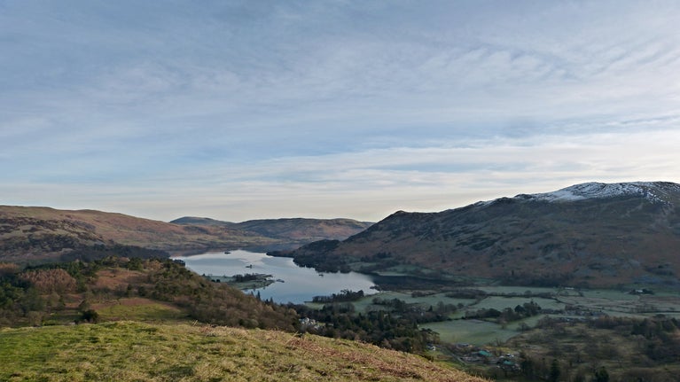 View of Ullswater and surrounding hills just after sunrise on an early spring day above the Patterdale Circular walking path. The sky is light blue with wispy clouds, the lake is still, and the fell tops have a dusting of snow. The valley floor is still in the shadow of the hills above.